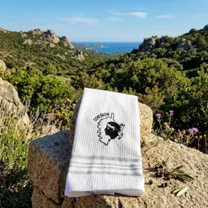 Torchon de cuisine blanc en nid d'abeille avec une broderie noire représentant la carte de la Corse, la Tête de Maure et l'inscription "Corsica". Le bas est orné de rayures grises. Le torchon est présenté plié