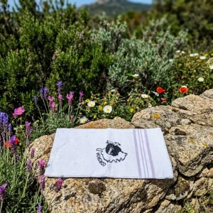 Torchon de cuisine blanc en nid d'abeille avec une broderie noire représentant la carte de la Corse, la Tête de Maure et l'inscription "Corsica". Le bas est orné de rayures coloris lavande. Le torchon est présenté plié